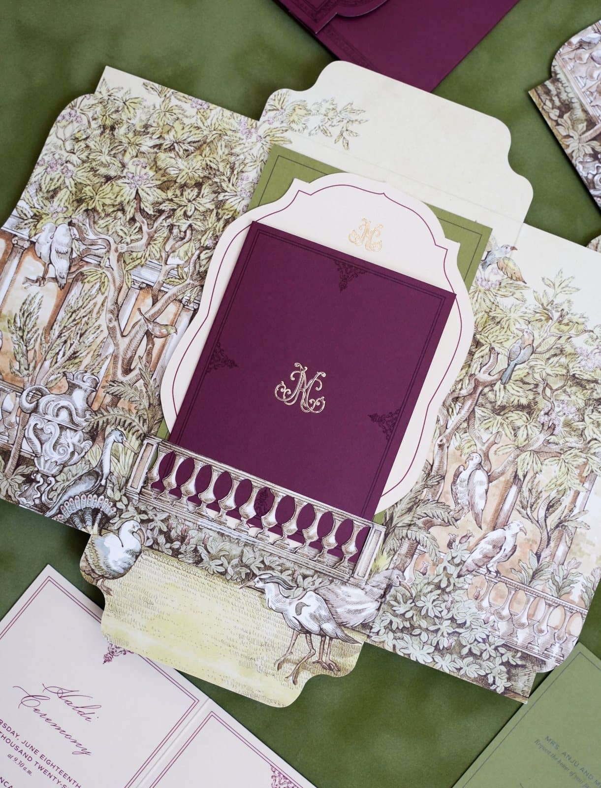 Open illustrated envelope with a die-cut balustrade framing a plum card with a gold foil monogram